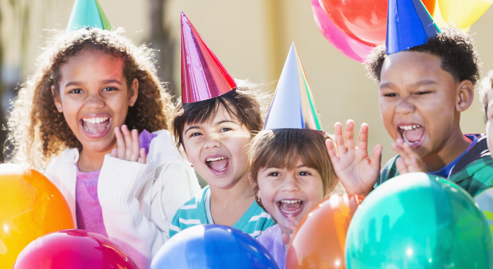 A group of four multi-ethnic children having fun at a birthday party laughing and waving, surrounded by colorful balloons and wearing party hats. They are mixed ages from 4 to 9 years old. The main focus is on the little girl wearing a pink hat.